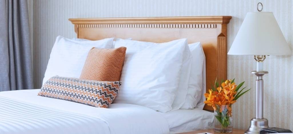 A neatly made hotel bed with white linens, decorative orange pillows and a bedside table holding a lamp and a vase of orange flowers.