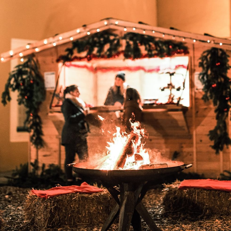 People gather around a fire pit near a decorated wooden stall with a vendor inside.