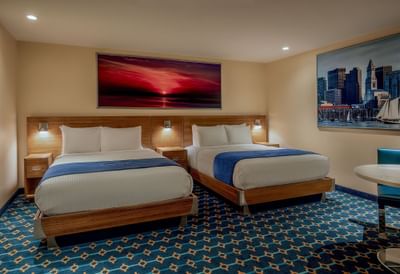 Two beds with sunset art above the headboard & city art on wall near a table in Double Queen Cabin at Harborside Inn Boston