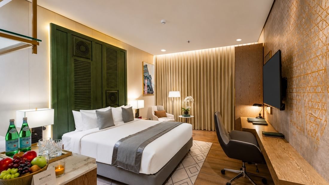 Bed with tall headboard facing the TV near work desk and glowing floor lamp in Accessible room at Naviti by Warwick Jeddah