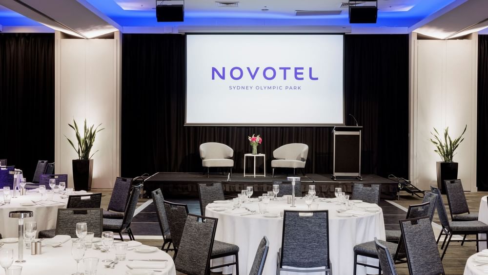 Banquet set-up with projector screen & stage featuring two plush seats in Freshwater Ballroom at Novotel Sydney Olympic Park