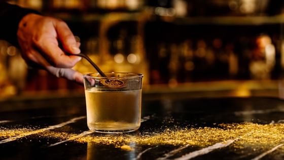 A bartender's hand stirs a cocktail surrounded by golden glitter on a countertop at The Kitchens