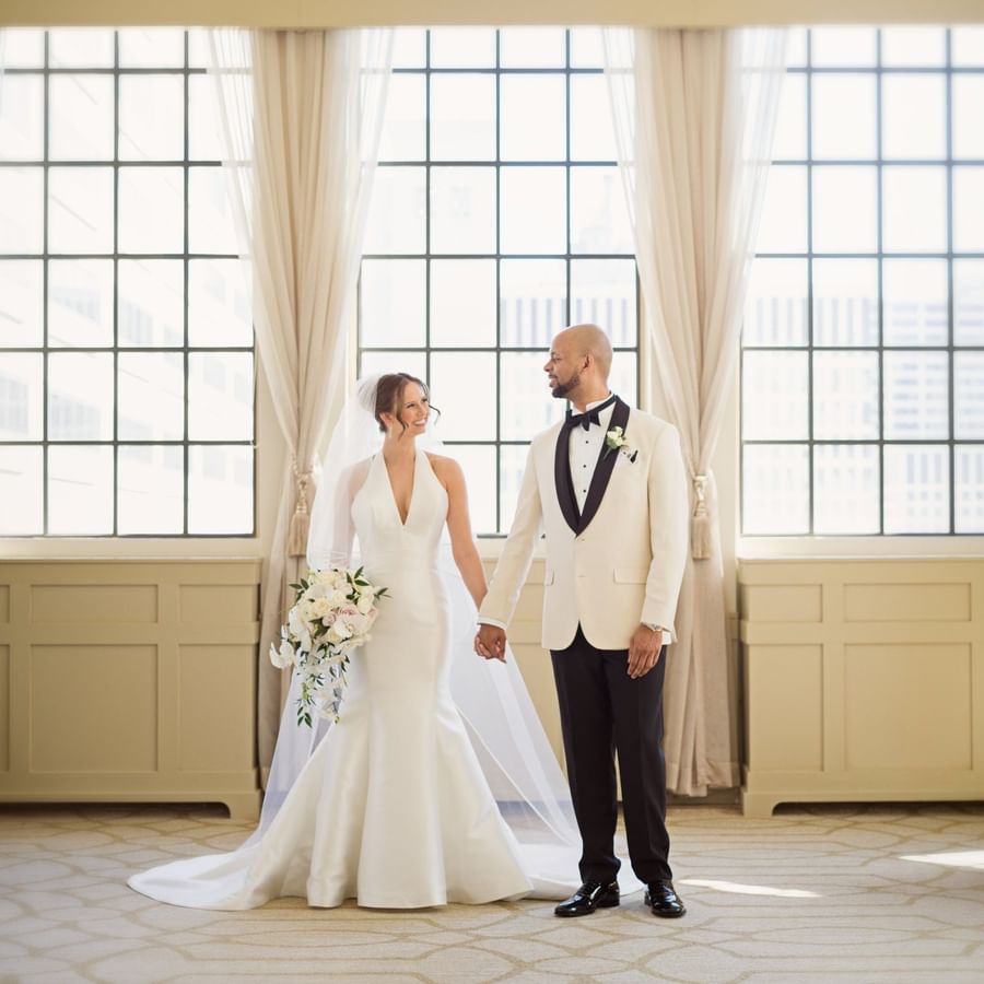 Bride in a white gown and groom in a tuxedo holding hands by a window at Warwick Allerton Chicago