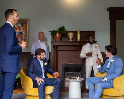 Five men in suits gather around a fireplace with drinks in a room with yellow chairs at The Ashbrook Hotel