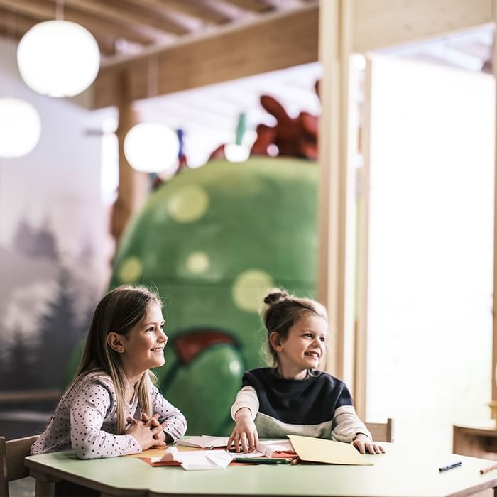 Zwei Mädchen sitzen an einem Tisch in einem Spielzimmer und lachen sich gegenseitig an.