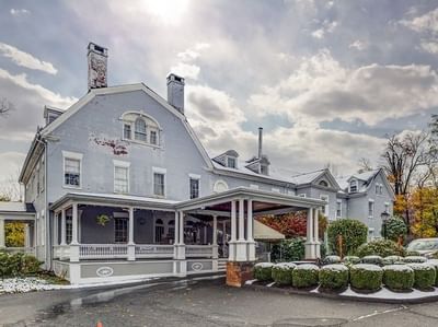 Exterior view of Simsbury 1820 House in winter, wraparound porch by a porte-cochere surrounded by snow-dusted shrubs