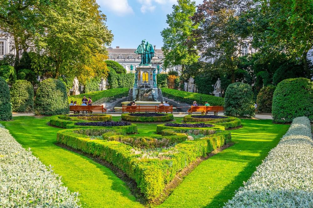 Garden with green hedges and a stone fountain placed by wooden benches, under trees at Warwick Grand Place Brussels