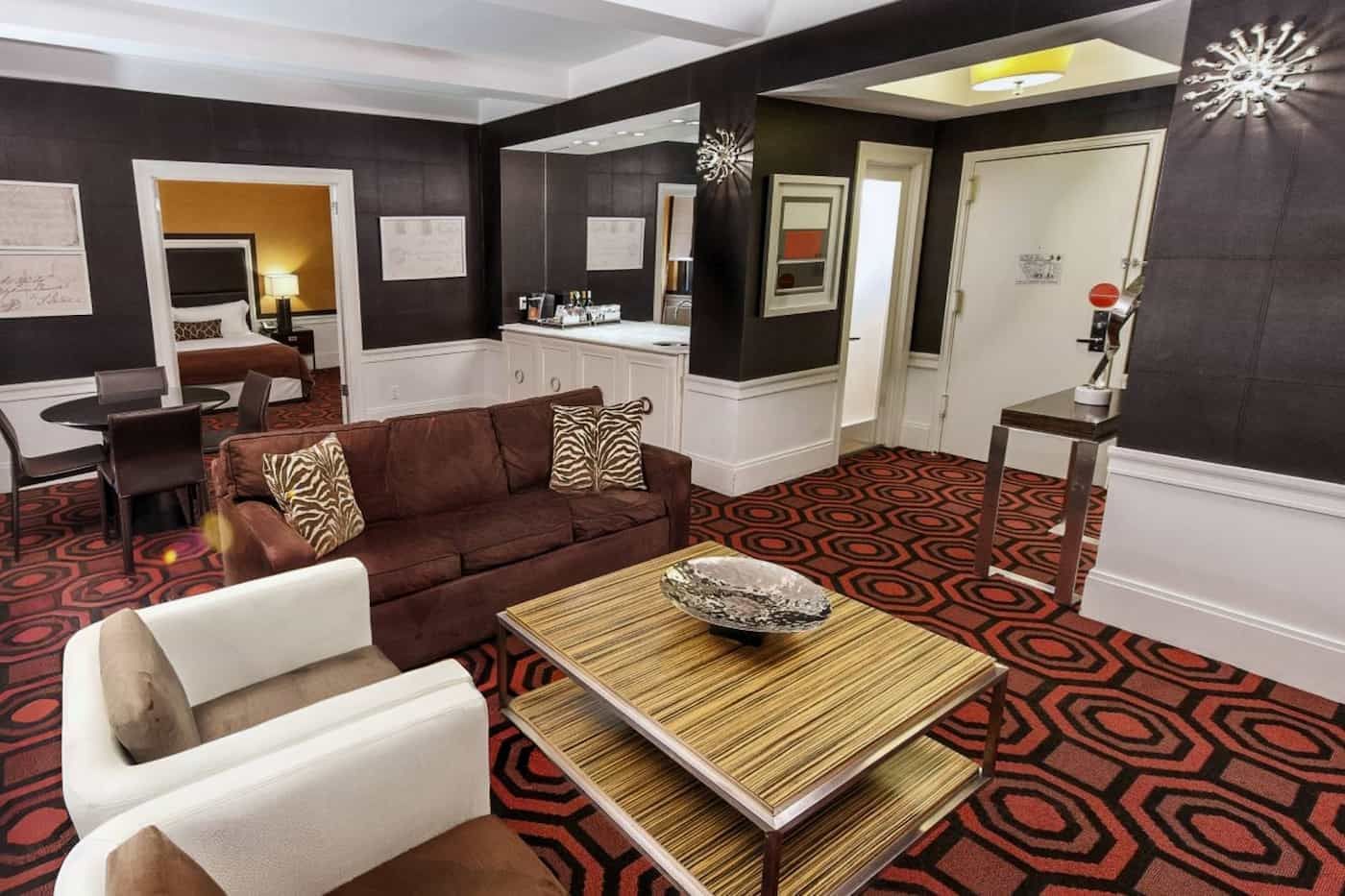 A wide view of a hotel suite's living area with a sofa, coffee table, armchairs, and a patterned carpet.