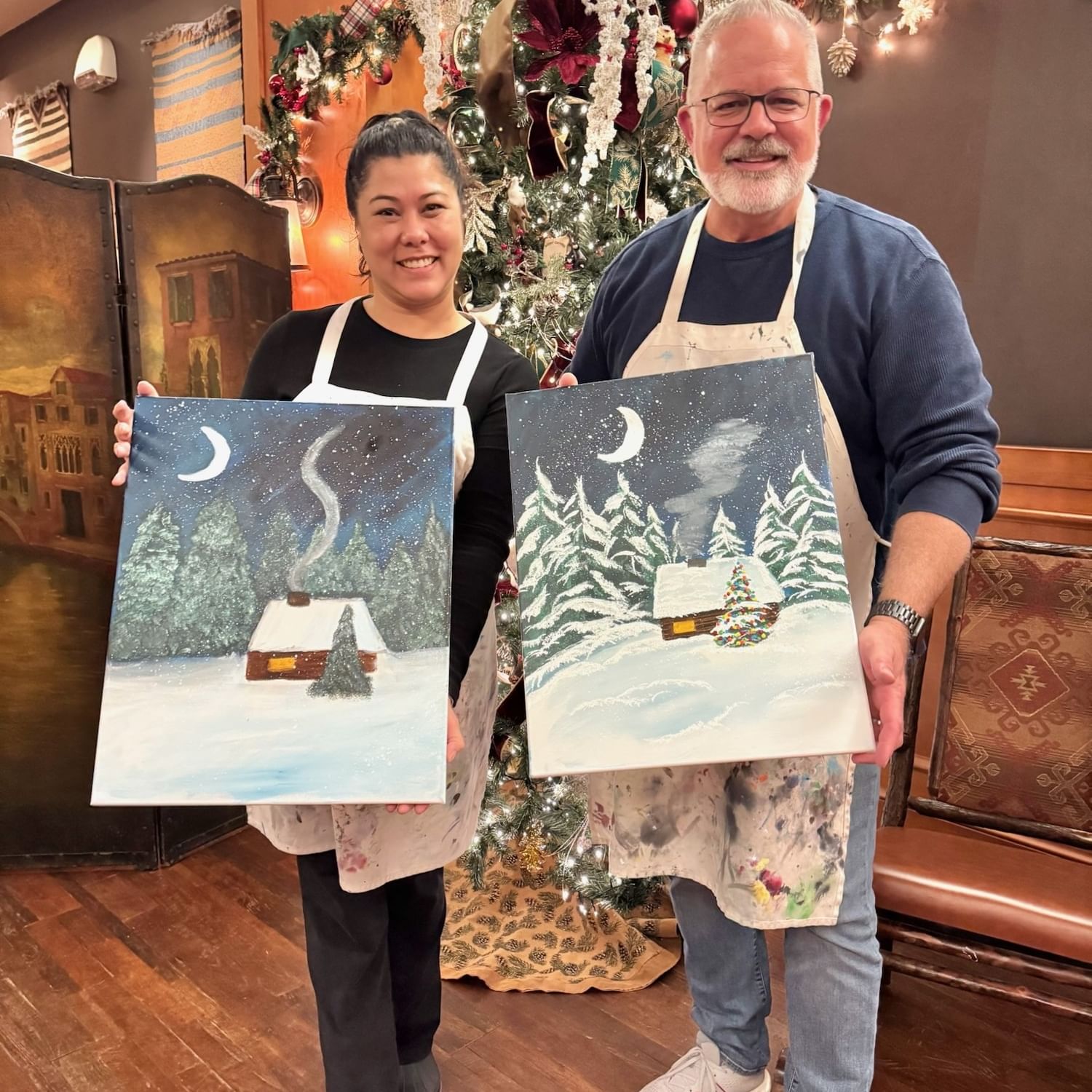 A man and woman with aprons hold their artwork in front of a decorated Christmas tree.