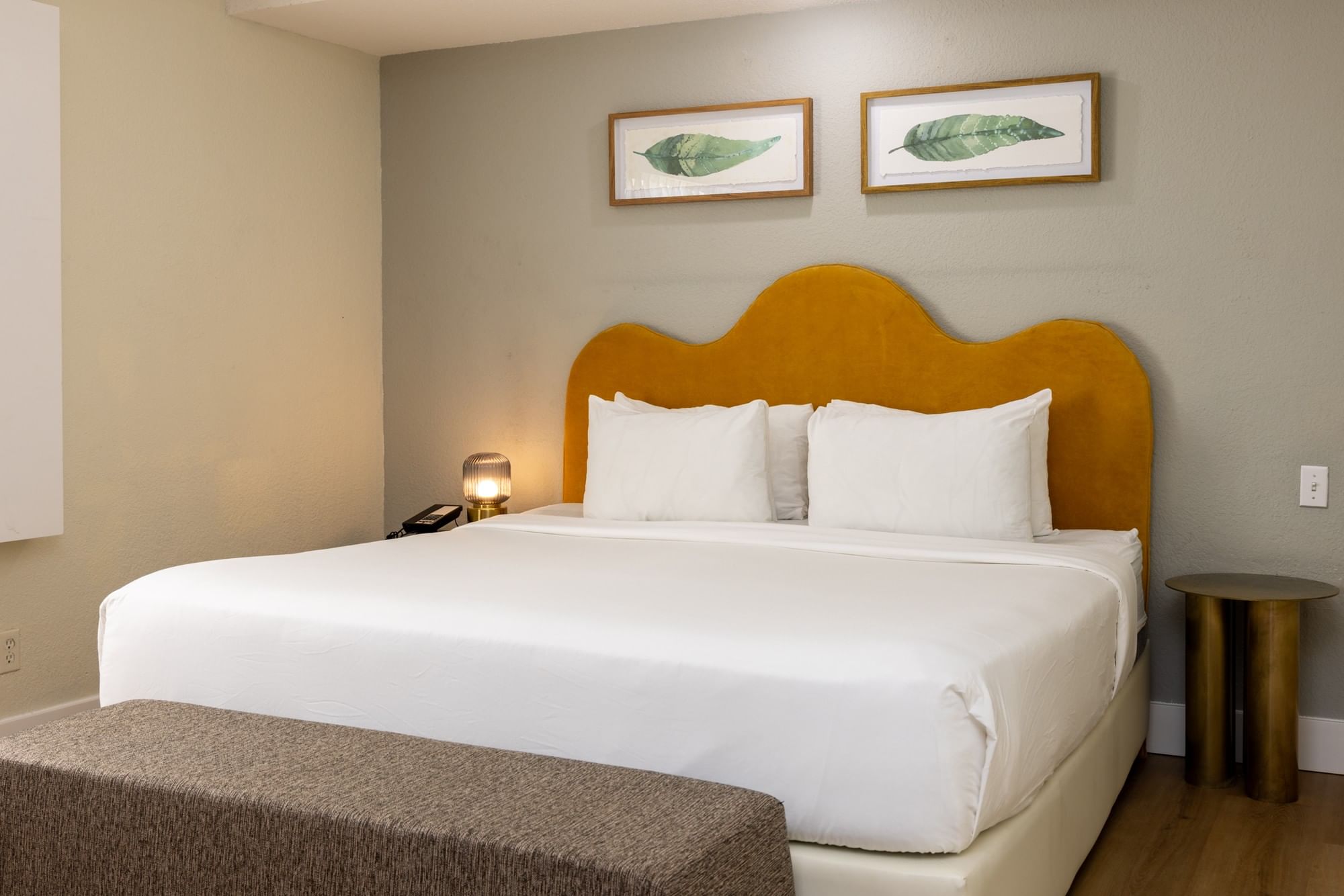 A hotel room with a king-sized bed, brown upholstered headboard, white bedding, and a brown bench at the foot of the bed.