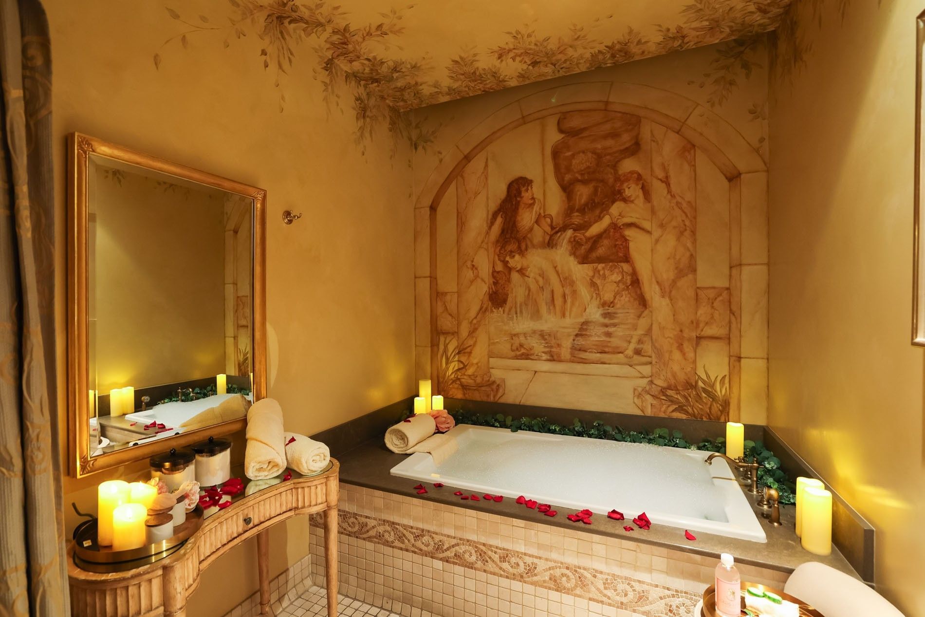 Luxury spa suite with a soaking tub, rose petals, glowing candles, and a classical stone mural at Kelly's Spa