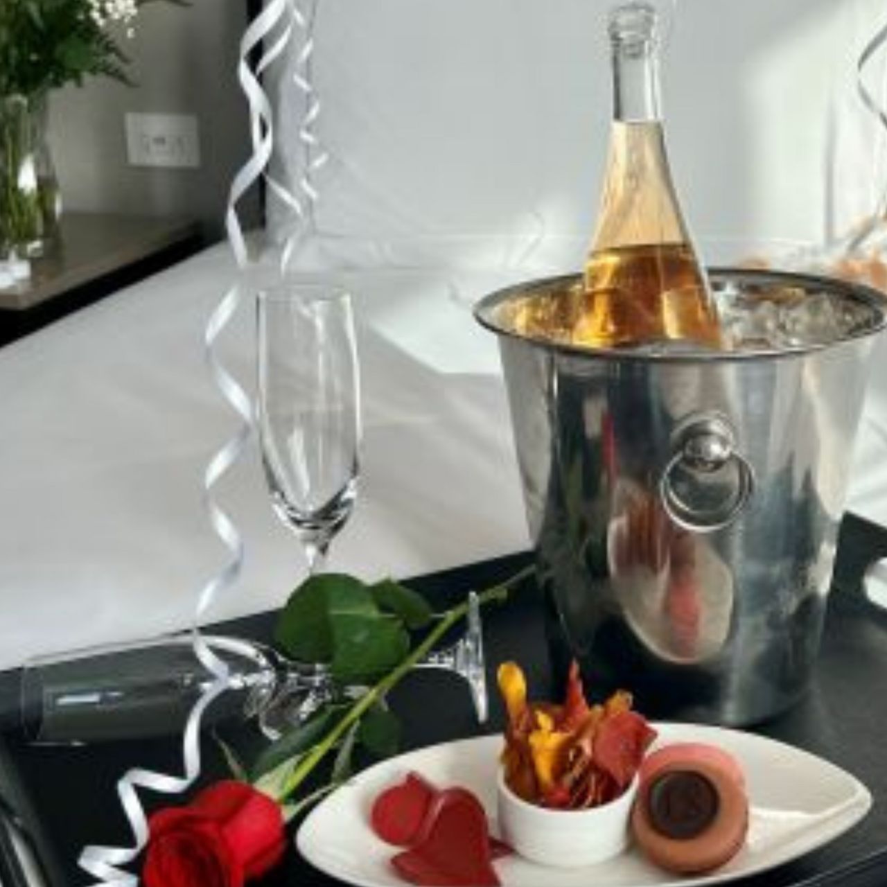 Sparkling wine in an ice bucket by a red rose, champagne glass and dessert plate arranged on the bed at Hotel X Toronto
