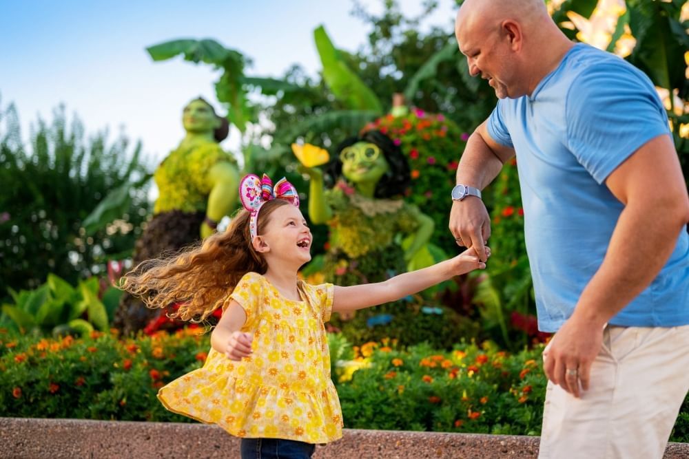 A father in a blue t-shirt twirls his daughter in a yellow shirt and colorful Minnie ears in front of a topiary display of characters from Encanto.  