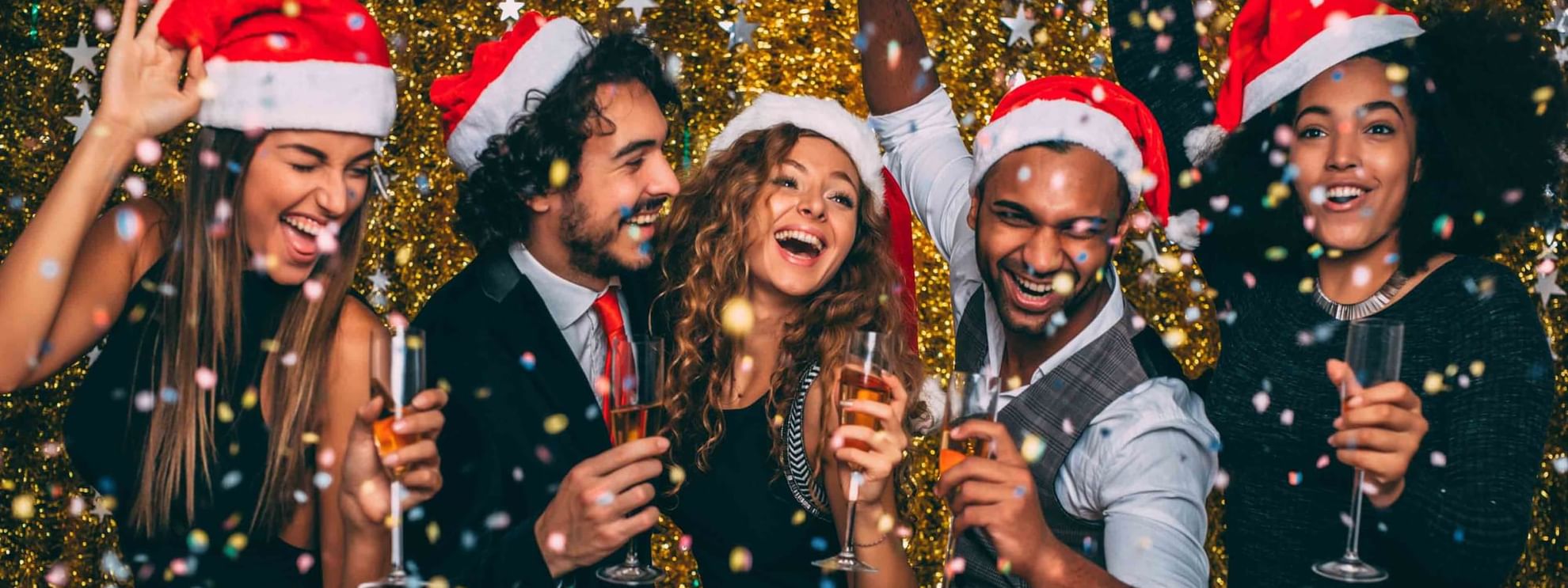 Five people in Santa hats holding wine glasses and celebrating in front of a glittery background.