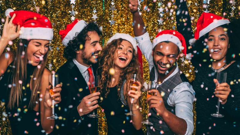 Five people in Santa hats holding wine glasses and celebrating in front of a glittery background.