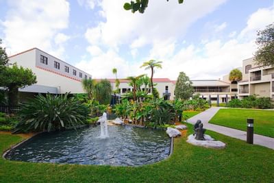 Garden pond with water fountain surrounded by tall palms and hotel balconies beside a paved walkway at Miami Lakes Hotel