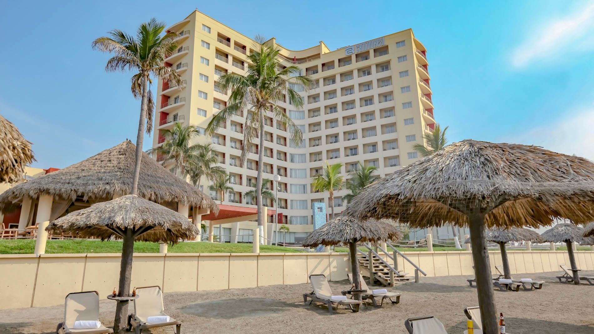 Tall yellow hotel exterior of Camino Real Veracruz viewed from the sandy beach with thatched palapa umbrellas