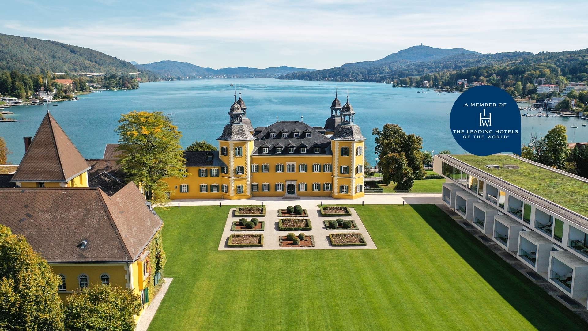 Luftaufnahme eines gelben Schlosses und grünen Rasens mit einem See und einem Hotel im Hintergrund.