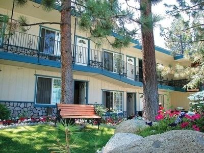 Exterior of a two-story building with blue trim, surrounded by pine trees and a lush garden area at Americana Village Suites