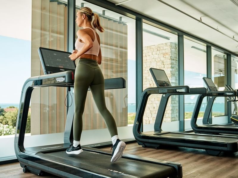 Eine Frau trainiert auf einem Laufband in einem Fitnessstudio mit großen Fenstern.