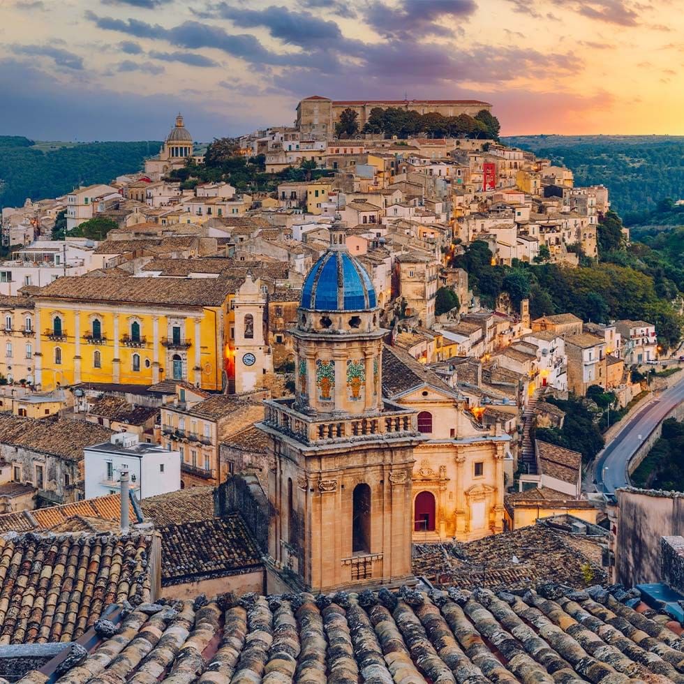 Vista aerea di un antico paese collinare con edifici storici e una cupola blu al tramonto.