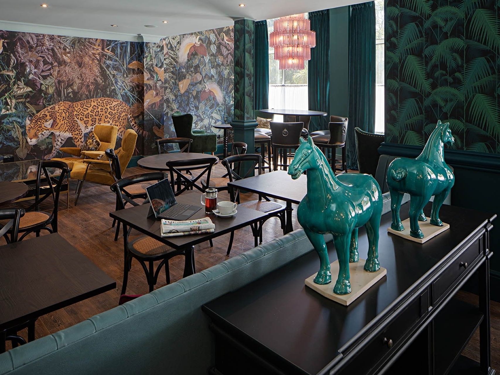 A restaurant with green horse statues on a table, tropical wall decor, and a chandelier above tables and chairs.