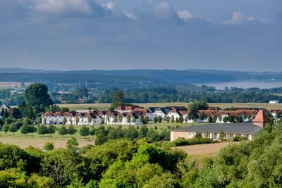 A rural landscape with a row of apartment houses, surrounded by trees and fields, under a partly cloudy sky.