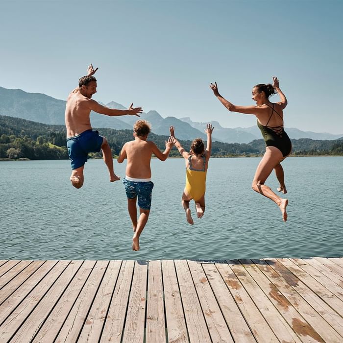 Vier Personen springen gleichzeitig von einem Steg in einen See, umgeben von Bergen.