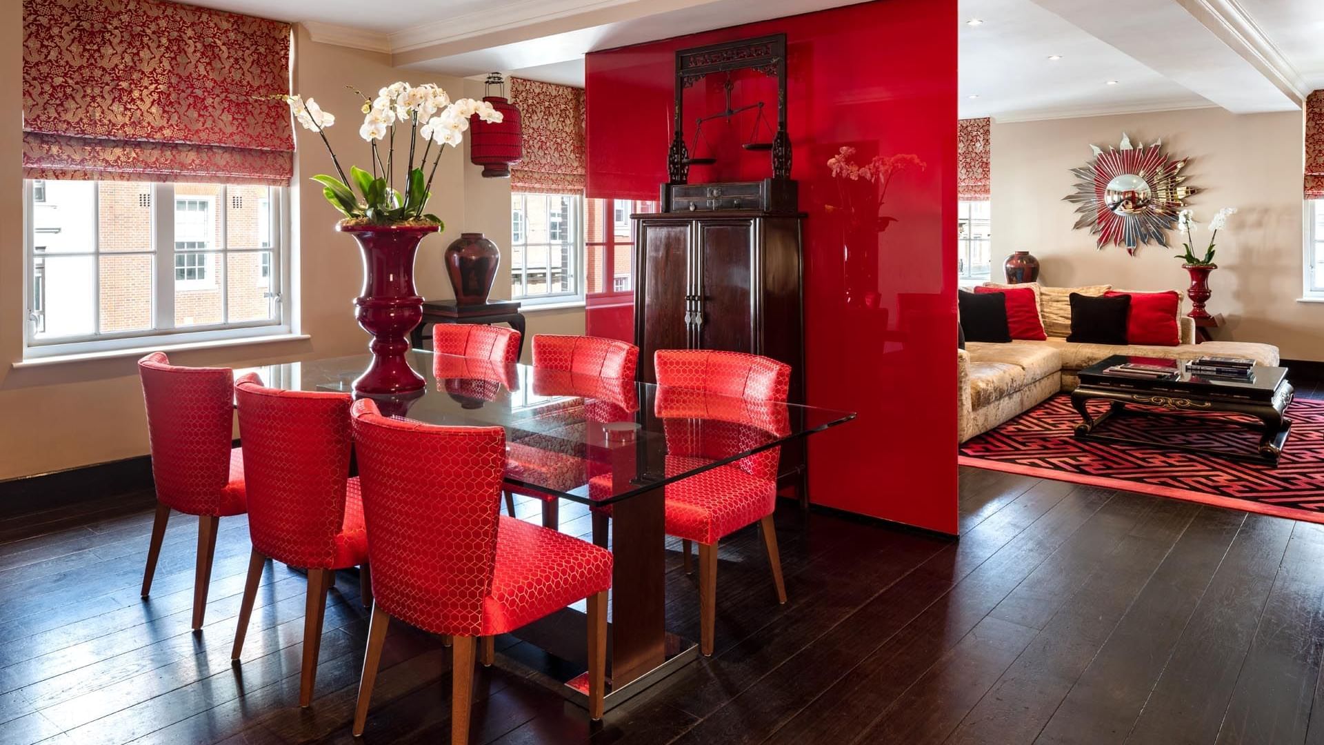 Luxurious dining room with red chairs, glass table, decorative vases, ornate wall art, and a plush beige sofa
