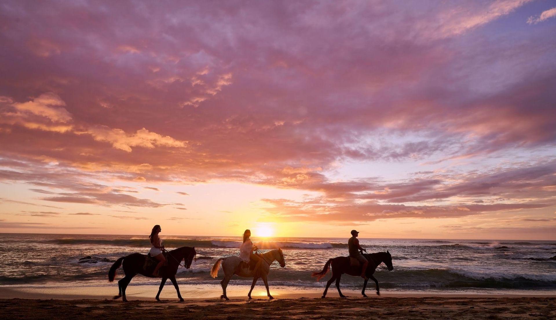 Three people riding horses on the beach during a vibrant purple and gold sunset near Cala Luna Boutique Hotel