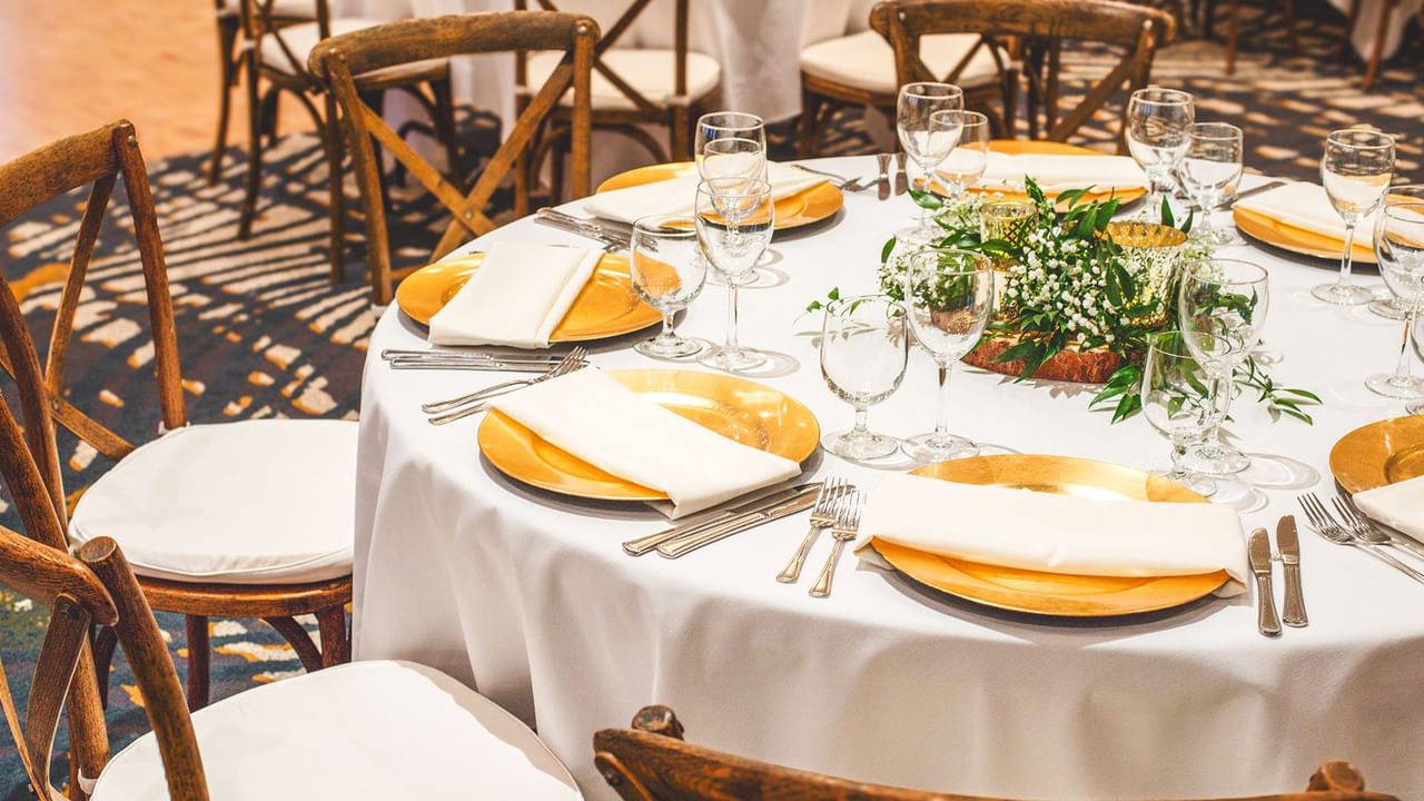 A round table with white tablecloth, gold plates, white napkins, cutlery, and wine glasses, surrounded by chairs.