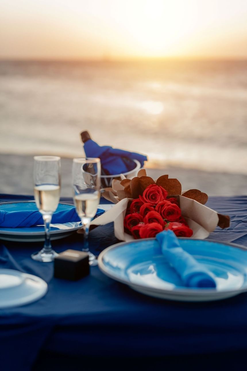 Beach table set with blue napkins, wine glasses, red roses, and a wine bottle at sunset.