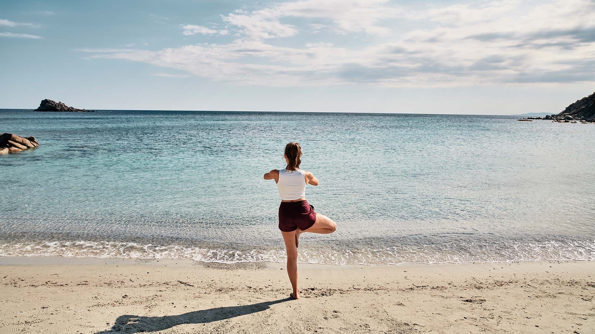 Woman doing yoga pose on sandy beach with ocean water and rocks in background.