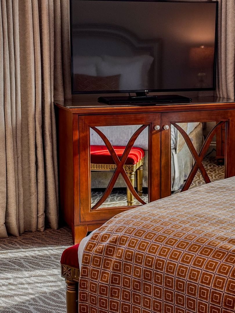 Large television on a wooden cabinet by a patterned bed under soft window light in Deluxe Suite at Warwick Melrose Dallas