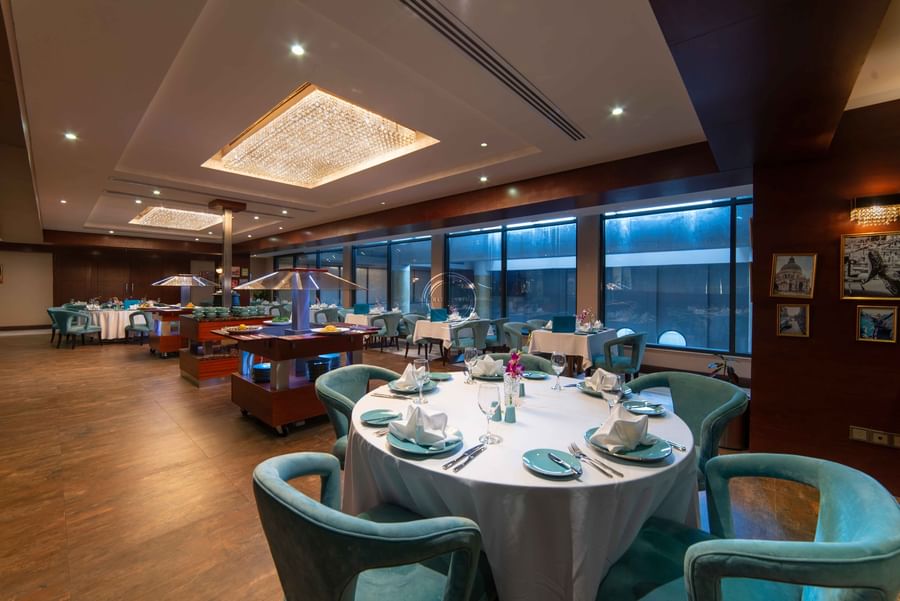 Teal velvet chairs set dining tables overlooking a pool and indoor garden atrium in Cigar Lounge at Warwick Al Khobar