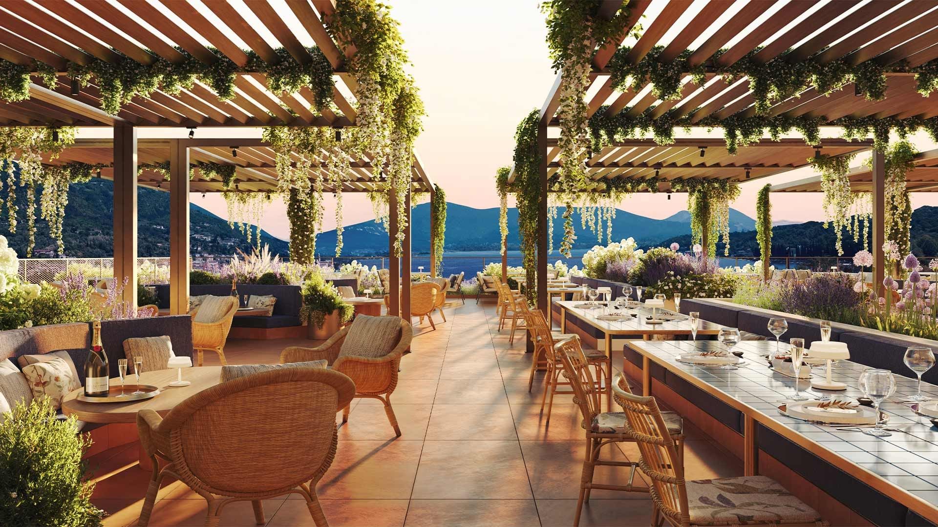 Terrazza di un ristorante con vista sul mare e montagne, sedie di vimini, tavoli imbanditi e pergolati decorati.