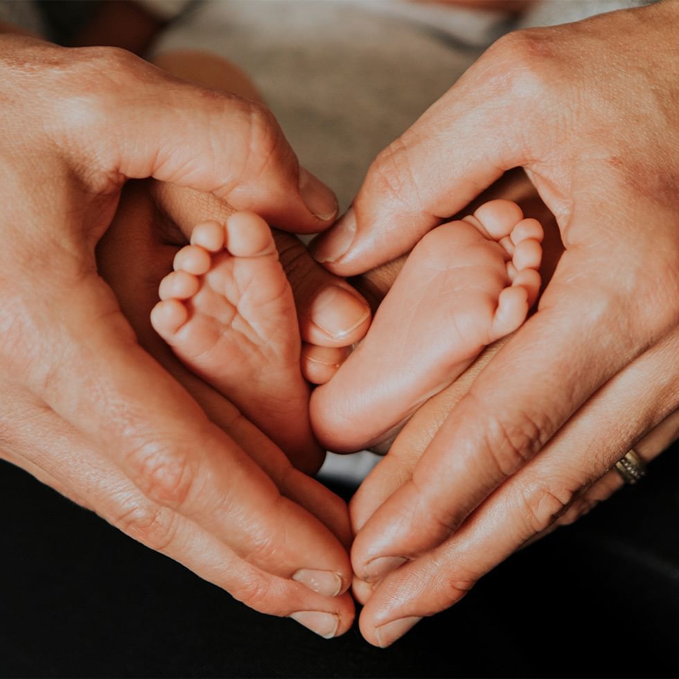 Zwei Hände halten in Herzform die nackten Füße eines Babys mit zehn Zehen.