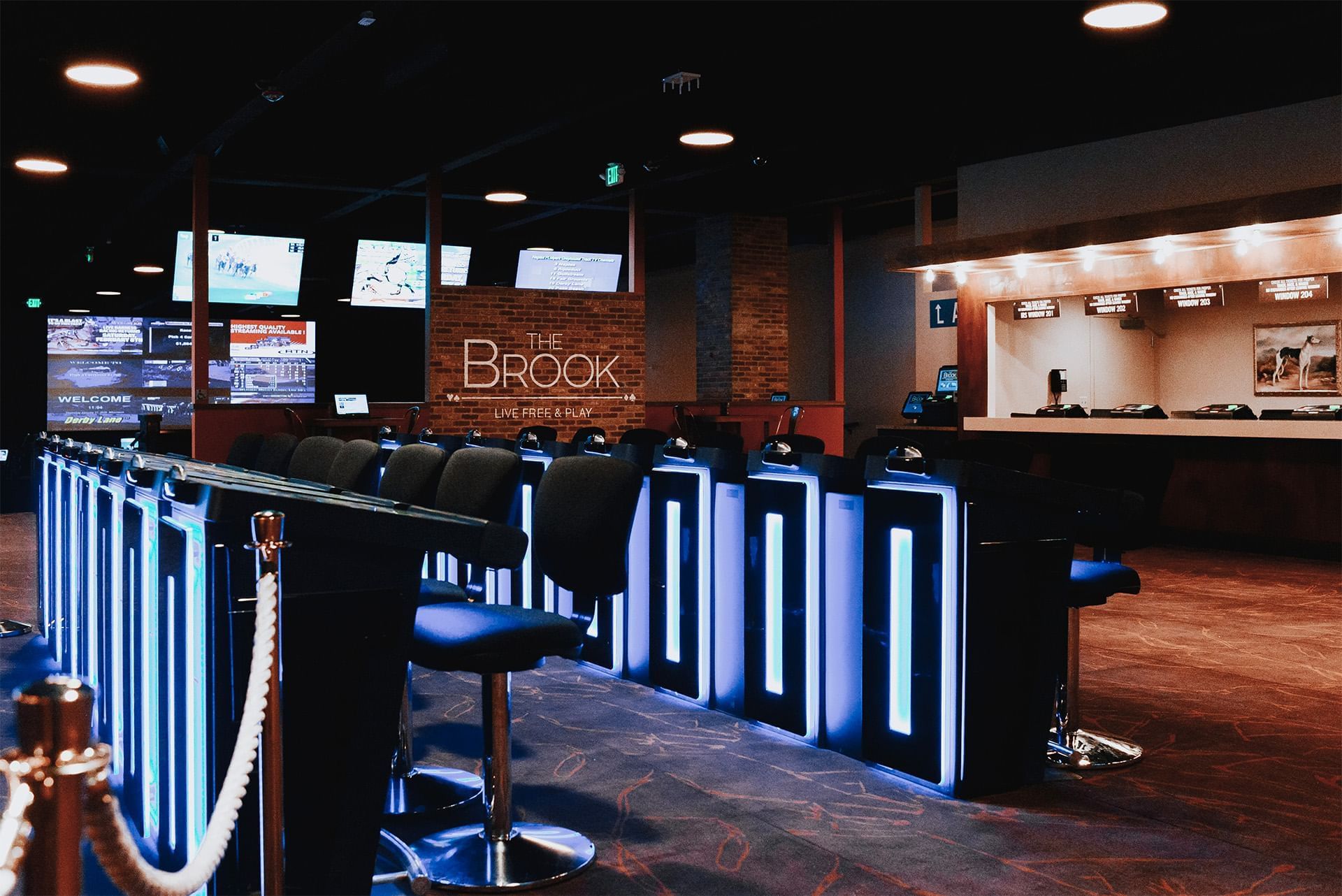 Stadium Games area with neon-lit betting terminals by cushioned stools, under TV screens beside signage at The Brook