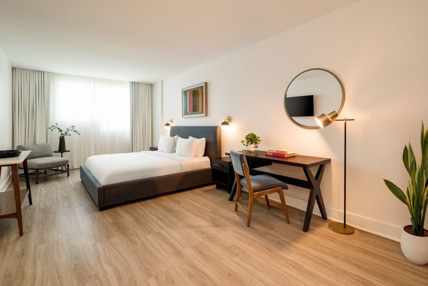 Modern hotel room with gray upholstered bed, wooden desk, round mirror, and light wood flooring 