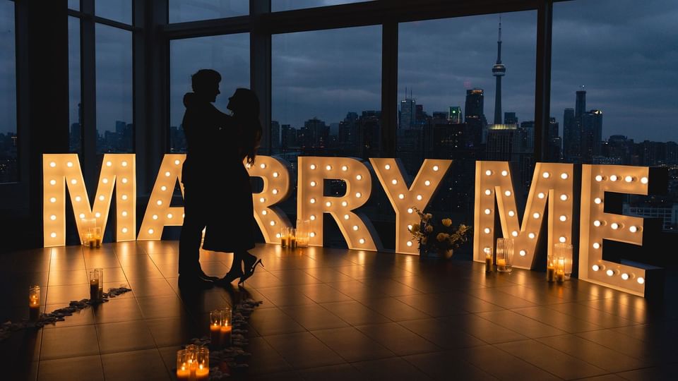 Couple silhouetted against Toronto skyline with illuminated MARRY ME marquee letters and candles at Hotel X Toronto