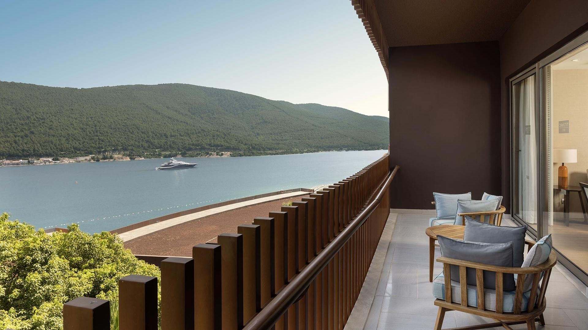 Deluxe Sea View Room's balcony with wooden furniture, overlooking a serene water body, surrounded by greenery and hills at Titanic Luxury Collection Bodrum.