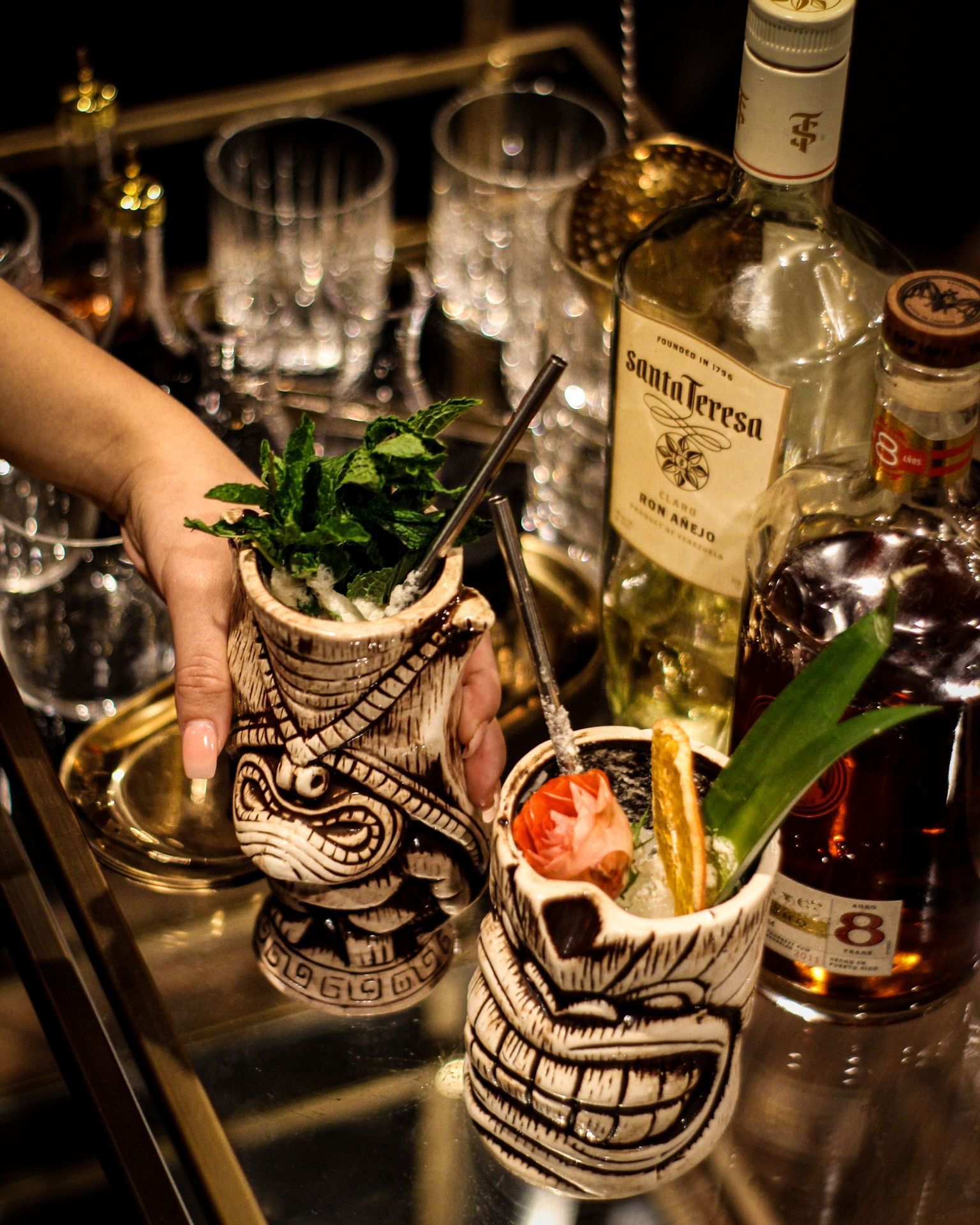 Two tiki glasses with straws and garnishes on a bar with bottles and glasses