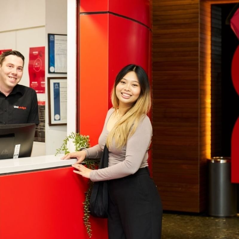 Two staff members smiling at a modern red and white reception desk with StudentLiving by UniLodge branding visible.