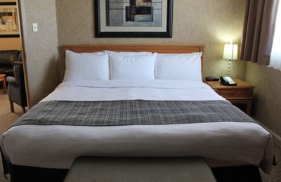 King bed with white pillows by a wooden nightstand and lamp, under framed art at Glenmore Inn & Convention Centre