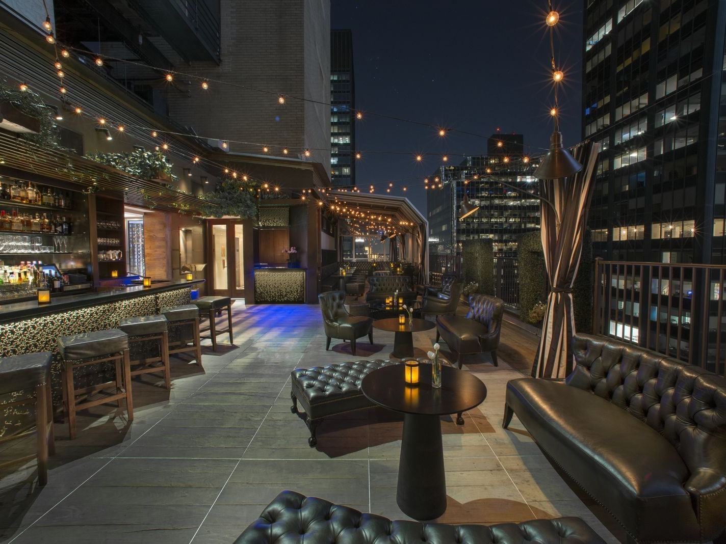Tufted leather sofas with round tables beside the rooftop bar illuminated by string lights in Upstairs at The Kimberly Hotel