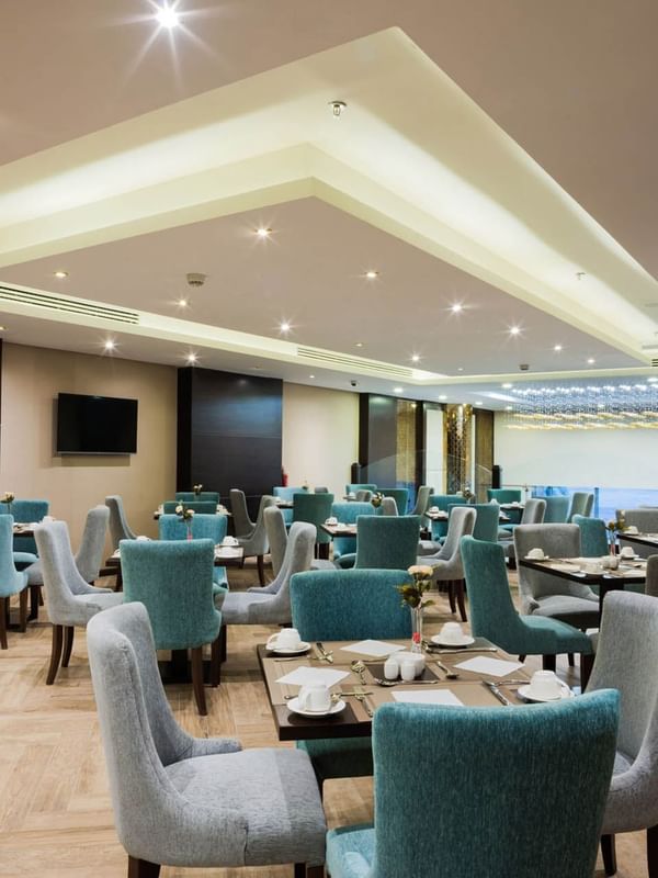 Blue armchairs by square dining tables under a recessed ceiling in Naviti Restaurant at Naviti By Warwick Dammam