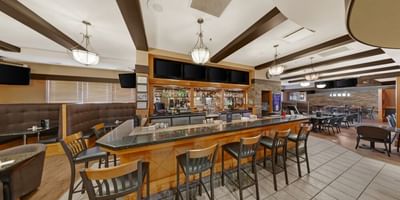 Wooden bar counter with black stools by mounted TVs, booth seating under pendant lights at Glenmore Inn & Convention Centre