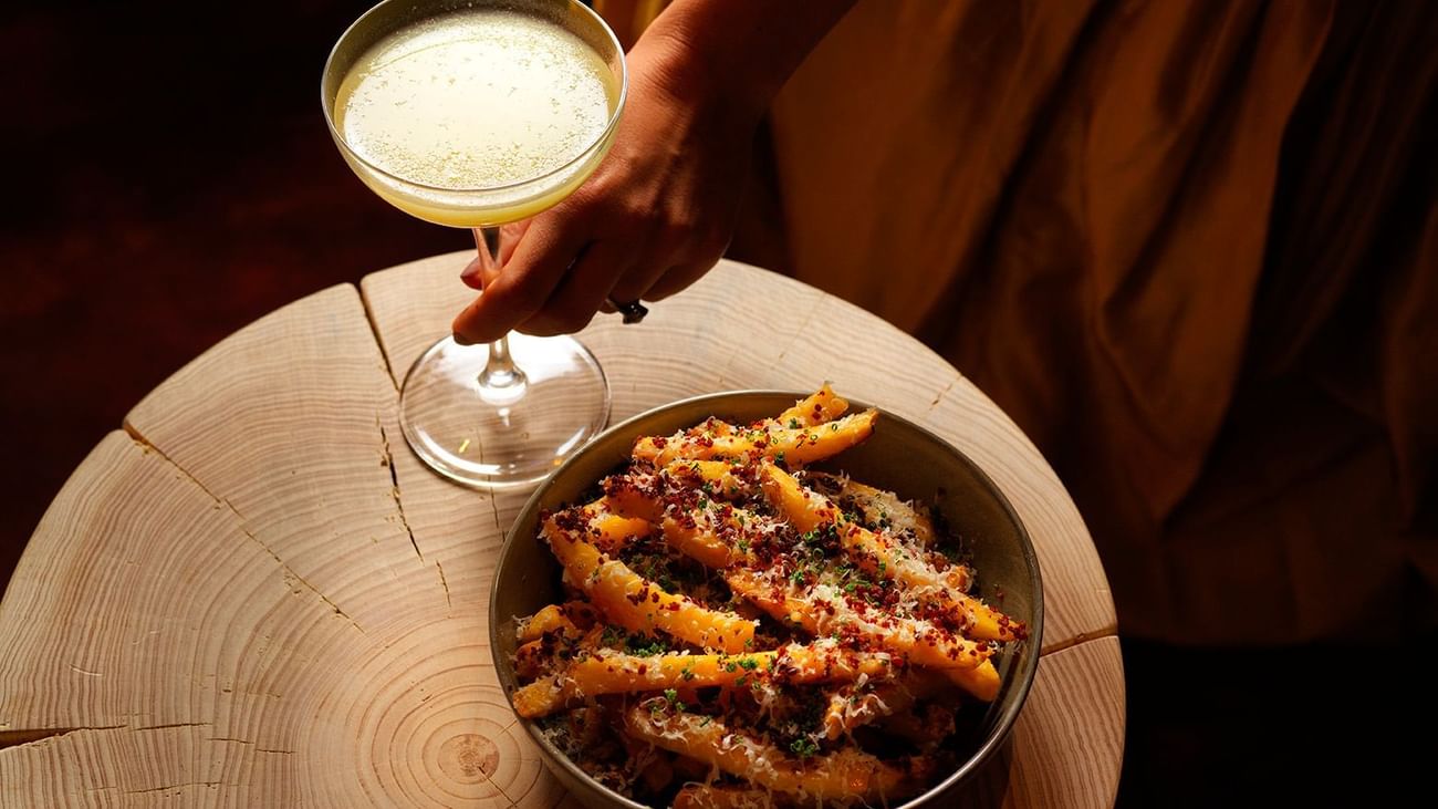 A person holds a cocktail glass near a bowl of fries on a wooden table.