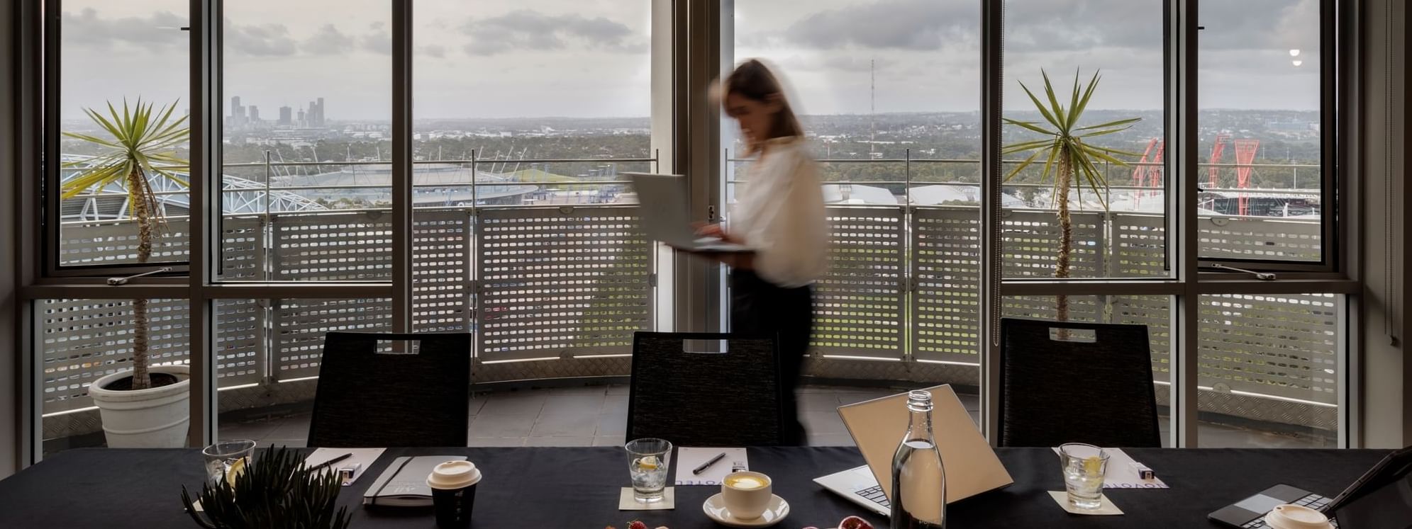 Table set-up with refreshments, laptop, and city views through large windows in The Lounge at Novotel Sydney Olympic Park