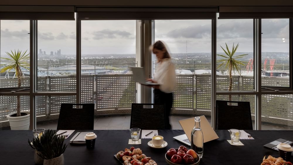 Table set-up with refreshments, laptop, and city views through large windows in The Lounge at Novotel Sydney Olympic Park